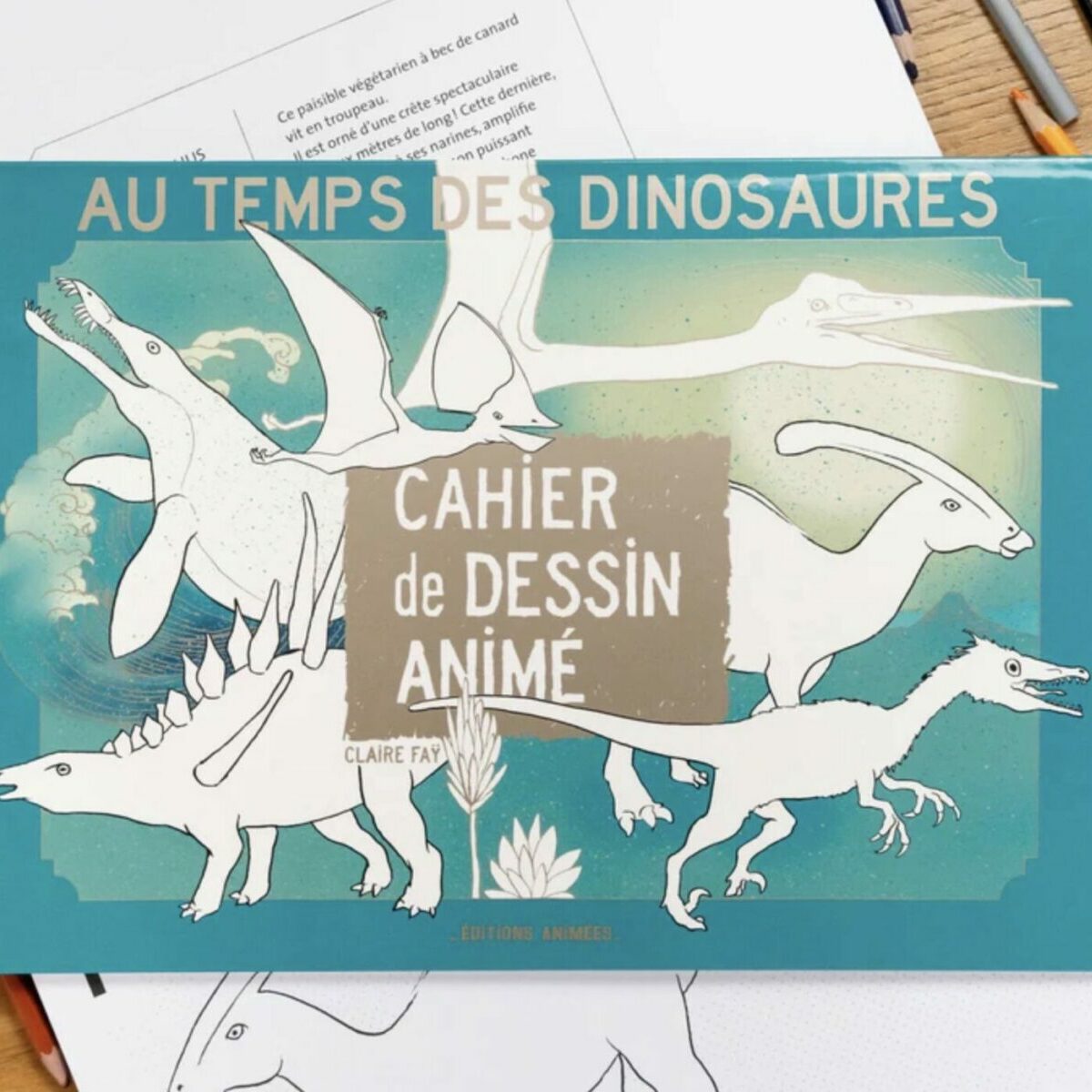 Couverture du Cahier de Dessin Animé Au Temps des Dinosaures BlinkBook posée sur un bureau avec crayons de couleur