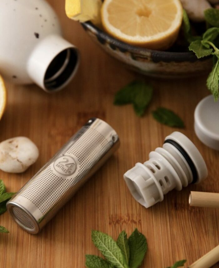 Photo en gros plan d'un infuseur à thé 24Bottles en acier inoxydable posé sur une table en bois, entouré de tranches de citron, de feuilles de menthe fraîche et d'une bouteille Clima blanche mouchetée.