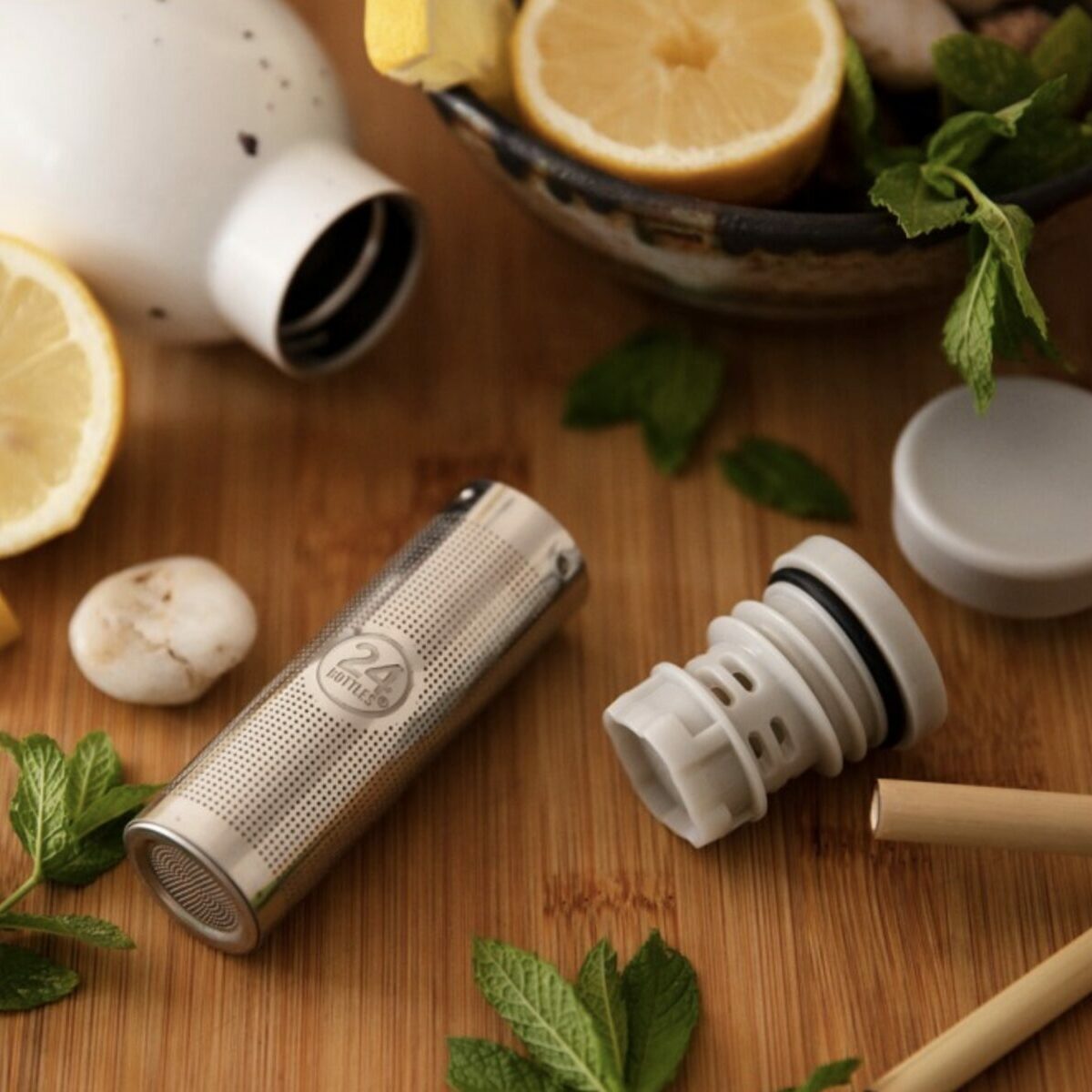 Photo en gros plan d'un infuseur à thé 24Bottles en acier inoxydable posé sur une table en bois, entouré de tranches de citron, de feuilles de menthe fraîche et d'une bouteille Clima blanche mouchetée.