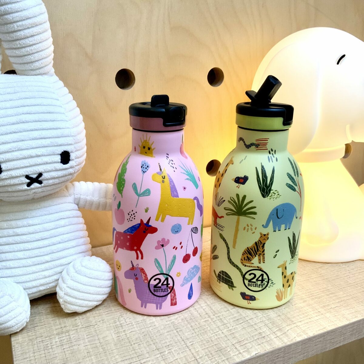 Deux gourdes 24Bottles Kids 300ml à motifs licornes et animaux de la jungle posées sur une étagère avec une peluche Miffy.