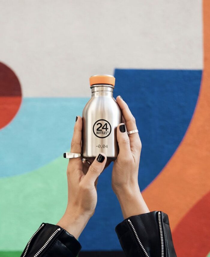 Mains tenant une petite gourde 24Bottles Urban Bottle 250ml en inox gris acier devant un mur de street art coloré.