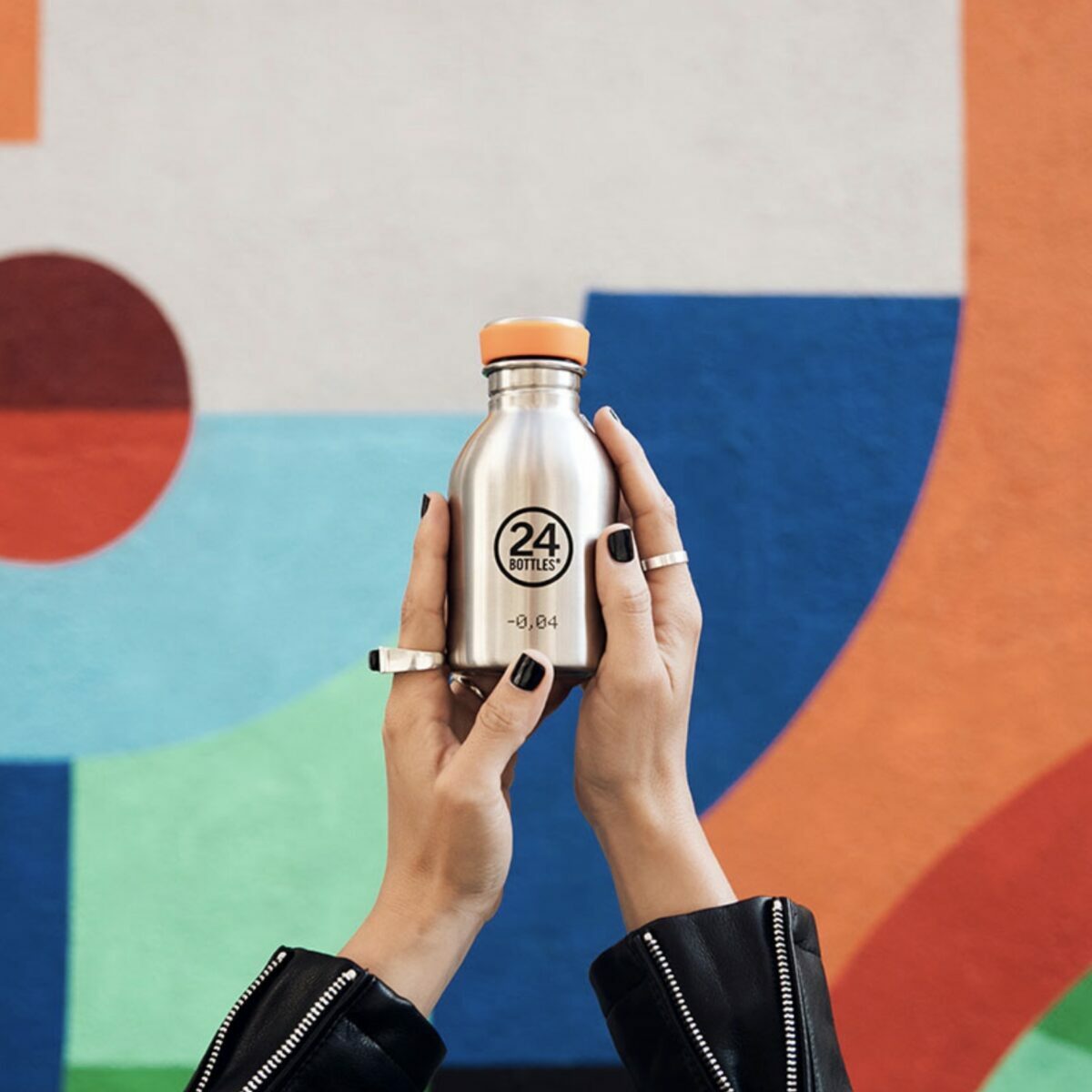 Mains tenant une petite gourde 24Bottles Urban Bottle 250ml en inox gris acier devant un mur de street art coloré.