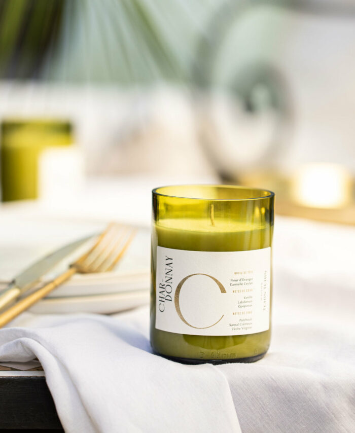 Bougie parfumée Chardonnay Maison Tchin Tchin en verre vert recyclé posée sur une table dressée avec nappe blanche et couverts dorés, d’autres bougies vertes allumées en arrière-plan, ambiance dîner élégant