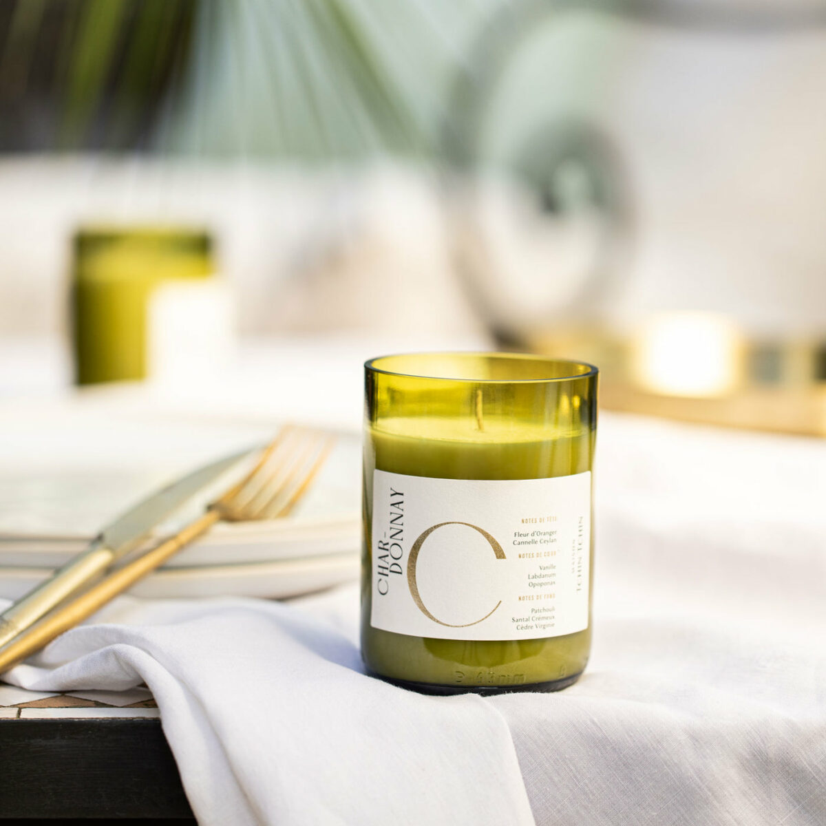 Bougie parfumée Chardonnay Maison Tchin Tchin en verre vert recyclé posée sur une table dressée avec nappe blanche et couverts dorés, d’autres bougies vertes allumées en arrière-plan, ambiance dîner élégant