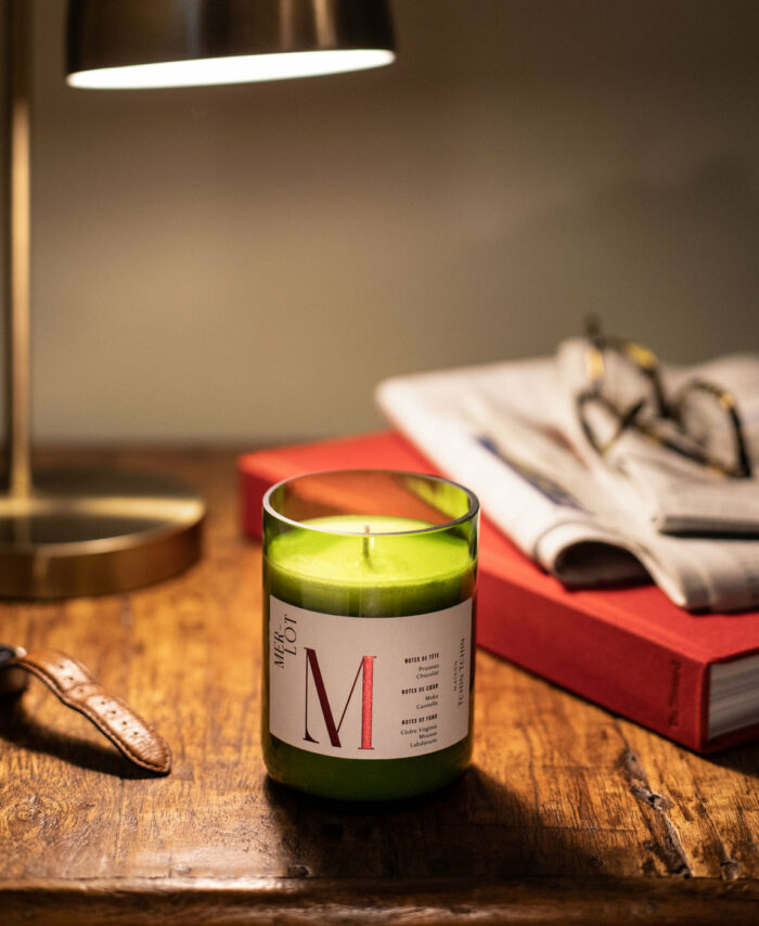 Bougie parfumée Merlot Maison Tchin Tchin en verre vert recyclé allumée sur un bureau en bois massif, à côté d’une lampe de bureau dorée, d’un livre rouge, d’un journal et d’une montre en cuir, ambiance cosy et tamisée