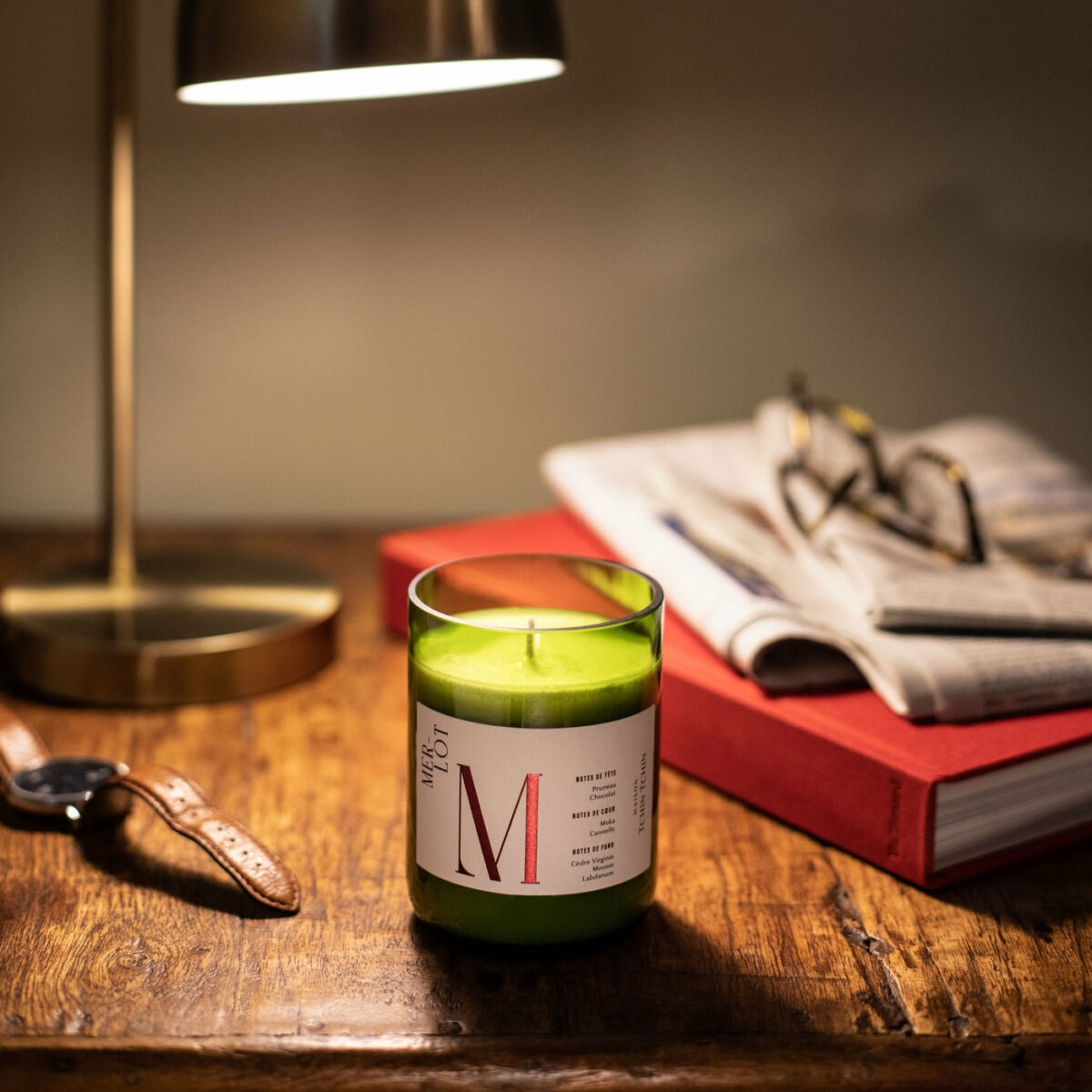 Bougie parfumée Merlot Maison Tchin Tchin en verre vert recyclé allumée sur un bureau en bois massif, à côté d’une lampe de bureau dorée, d’un livre rouge, d’un journal et d’une montre en cuir, ambiance cosy et tamisée