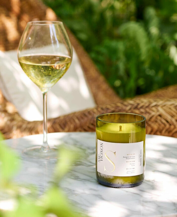Bougie parfumée Sauvignon Maison Tchin Tchin en verre vert recyclé posée sur une table en marbre blanc, à côté d’un verre de vin blanc, dans un jardin ensoleillé avec fauteuil en rotin et végétation verte en arrière-plan
