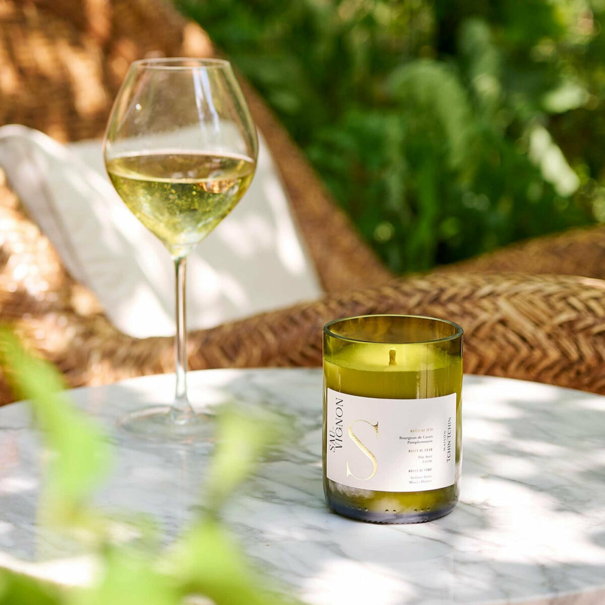 Bougie parfumée Sauvignon Maison Tchin Tchin en verre vert recyclé posée sur une table en marbre blanc, à côté d’un verre de vin blanc, dans un jardin ensoleillé avec fauteuil en rotin et végétation verte en arrière-plan