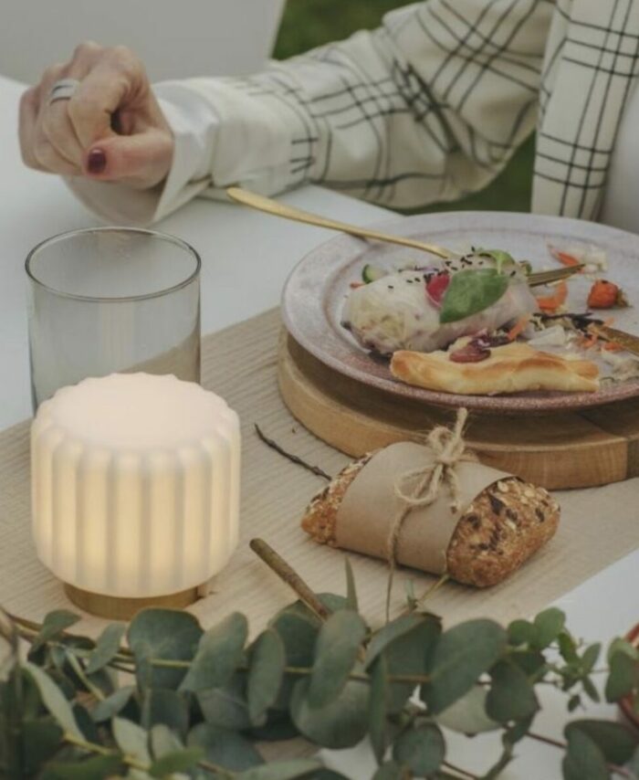 Mini lampe Dentelle Atelier Pierre allumée sur une table de repas en extérieur, abat-jour strié en verre dépoli blanc et base laiton doré, à côté d’un verre d’eau et d’une assiette avec couverts dorés, branches d’eucalyptus au premier plan