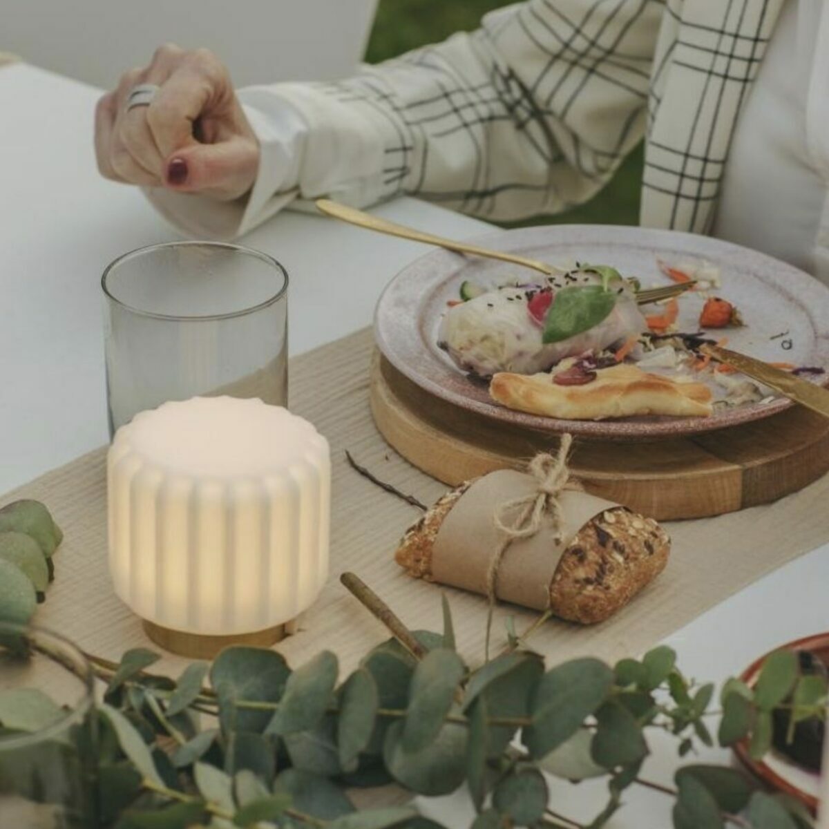 Mini lampe Dentelle Atelier Pierre allumée sur une table de repas en extérieur, abat-jour strié en verre dépoli blanc et base laiton doré, à côté d’un verre d’eau et d’une assiette avec couverts dorés, branches d’eucalyptus au premier plan
