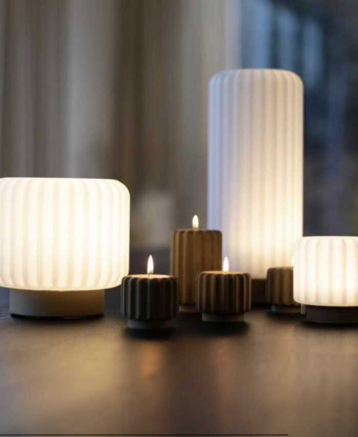 Composition intérieure de lampes Dentelle Atelier Pierre en format medium et mini allumées sur une table en bois sombre, associées à des bougeoirs striés dorés avec bougies allumées, lumière naturelle de fenêtre en arrière-plan avec rideaux blancs