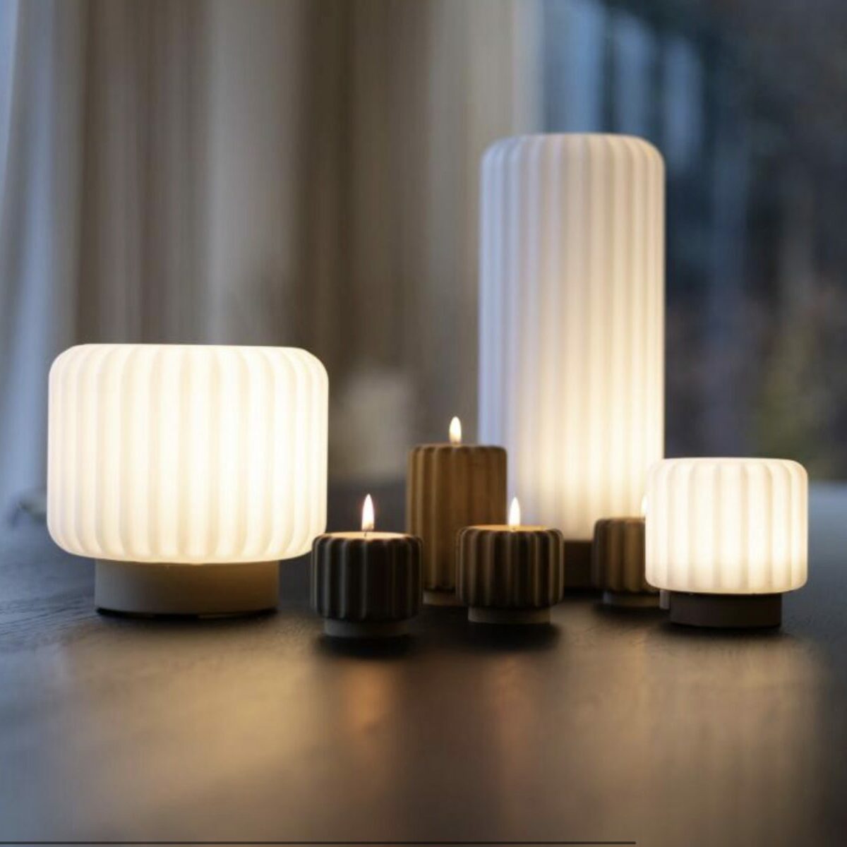 Composition intérieure de lampes Dentelle Atelier Pierre en format medium et mini allumées sur une table en bois sombre, associées à des bougeoirs striés dorés avec bougies allumées, lumière naturelle de fenêtre en arrière-plan avec rideaux blancs