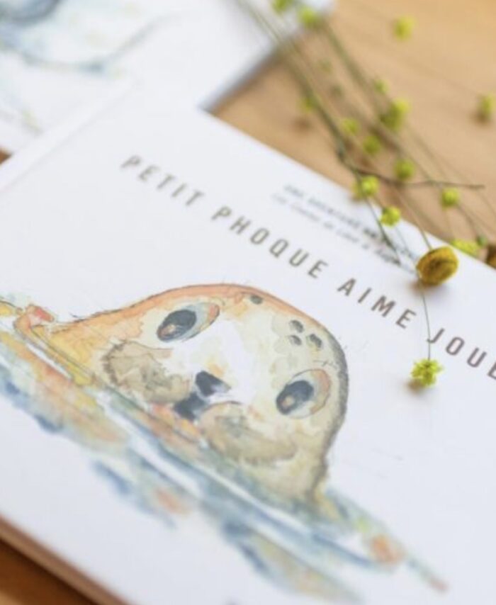 Livre jeunesse Petit Phoque aime jouer des Contes de Léon et Augustine posé sur une table en bois clair avec fleurs séchées jaunes, couverture blanche avec illustration aquarelle d’un phoque aux grands yeux émergeant de l’eau