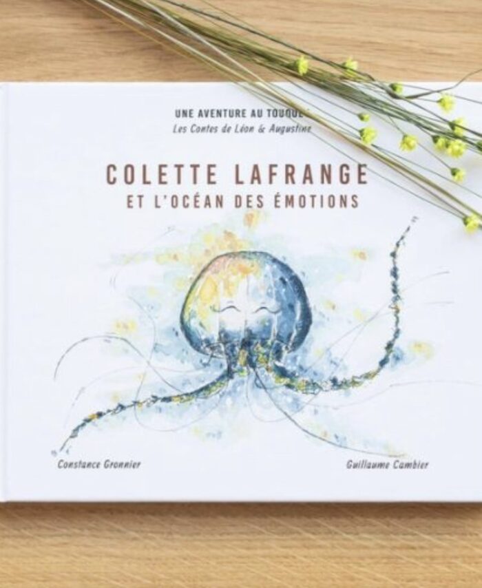 Livre jeunesse Colette Lafrange et l’Océan des Émotions des Contes de Léon et Augustine posé à plat sur une table en bois clair avec tiges de fleurs séchées jaunes, couverture blanche avec illustration aquarelle d’une grande méduse bleue aux longs filaments