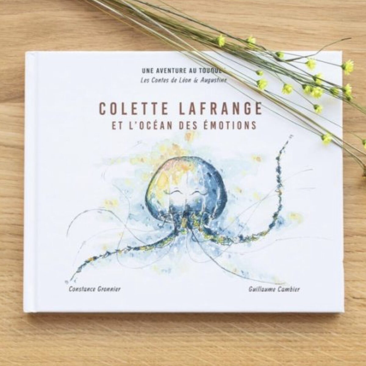 Livre jeunesse Colette Lafrange et l’Océan des Émotions des Contes de Léon et Augustine posé à plat sur une table en bois clair avec tiges de fleurs séchées jaunes, couverture blanche avec illustration aquarelle d’une grande méduse bleue aux longs filaments