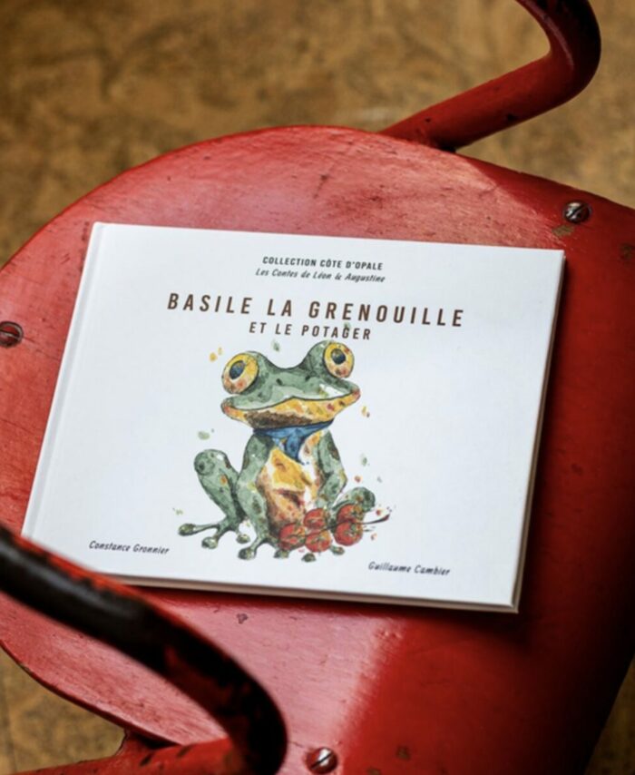 Livre jeunesse Basile la Grenouille et le Potager des Contes de Léon et Augustine posé sur une chaise en métal rouge vieilli, couverture blanche avec illustration aquarelle de Basile la grenouille en chapeau bleu tenant des tomates rouges